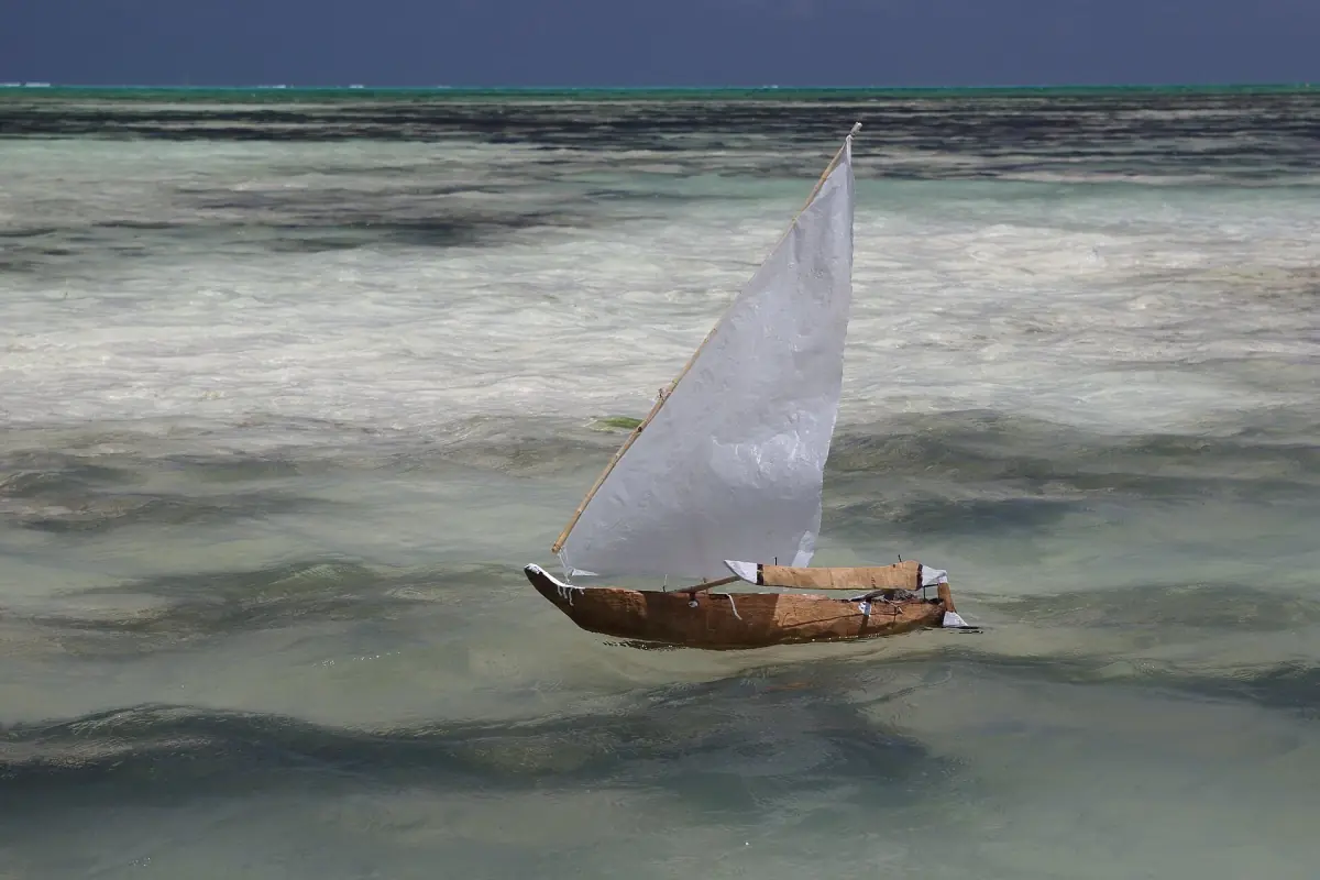 Hand-built model dhow with a white sail floating in shallow water at Jambiani